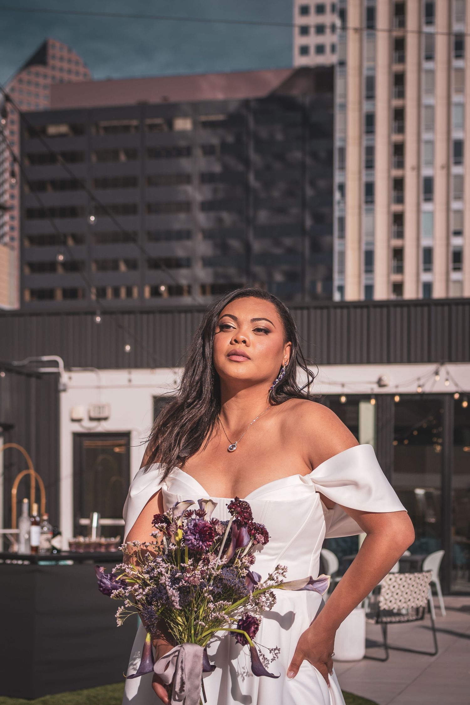 Elegant bride in white off-shoulder dress holding purple flowers against urban backdrop downtown Denver.