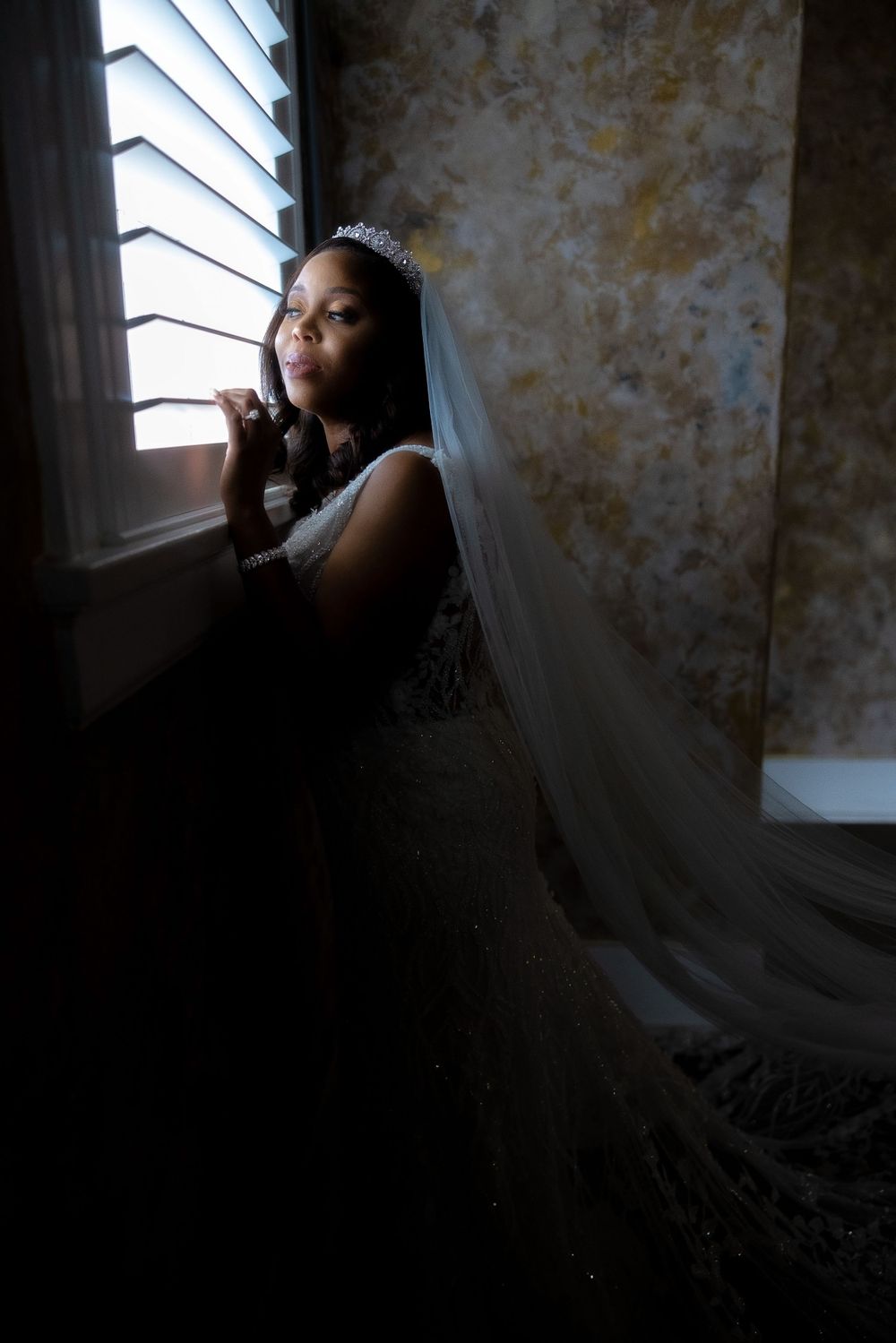 Artistic silhouette of bride in lace gown against textured gold wall and window light.