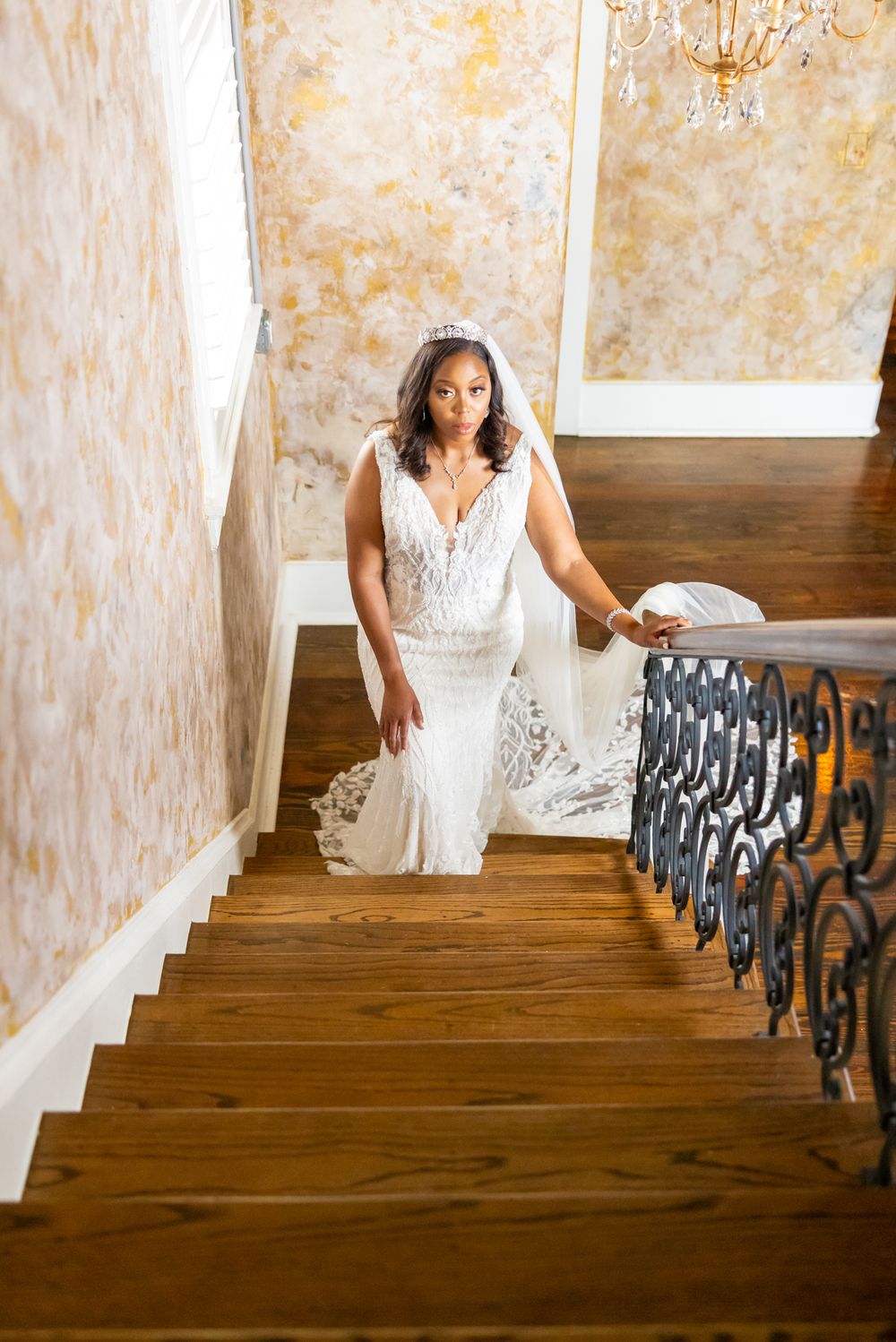 Bride in fitted lace gown ascending wooden staircase with ornate iron railing.