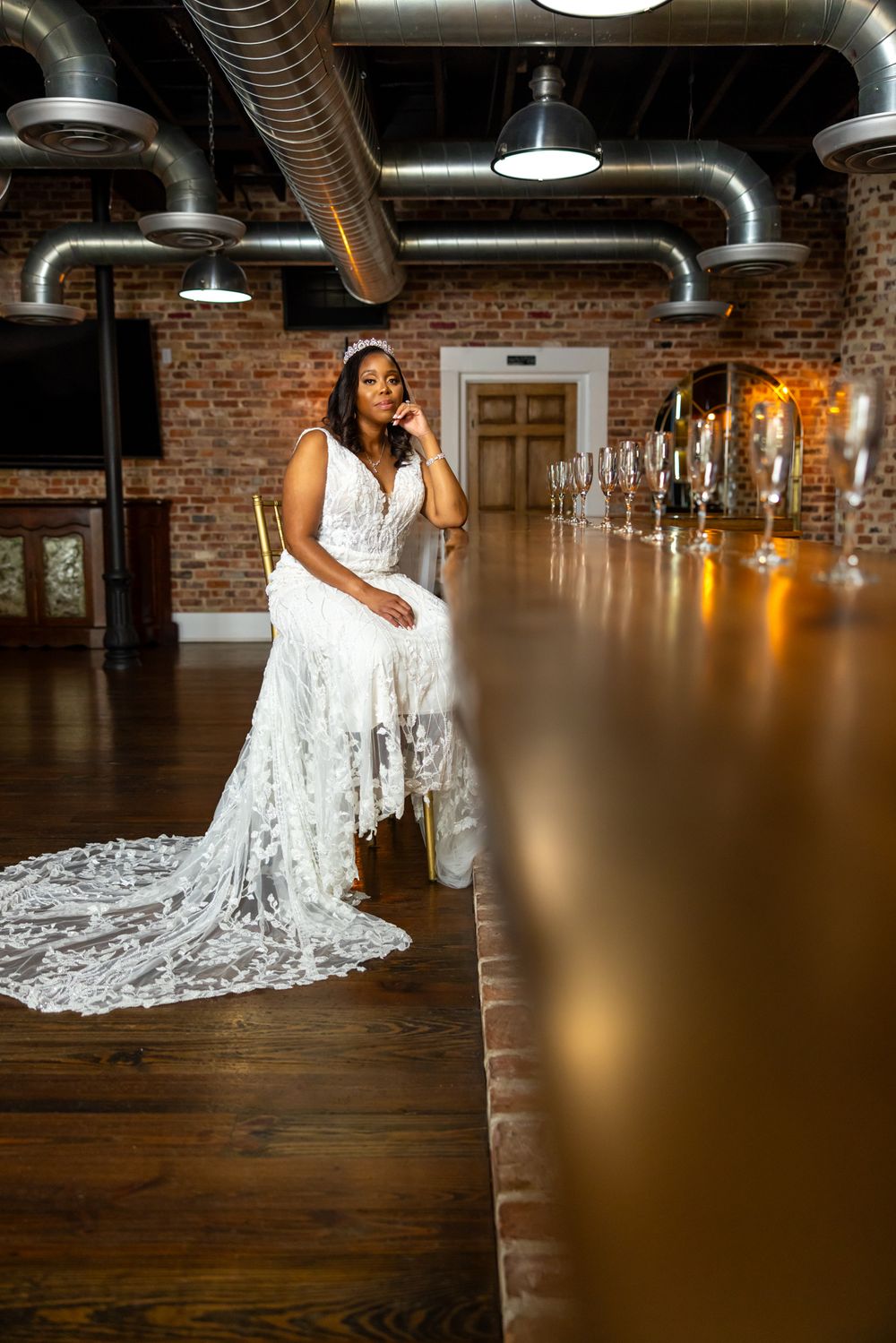 Bride seated at long wooden table in industrial space with exposed ductwork and brick walls.