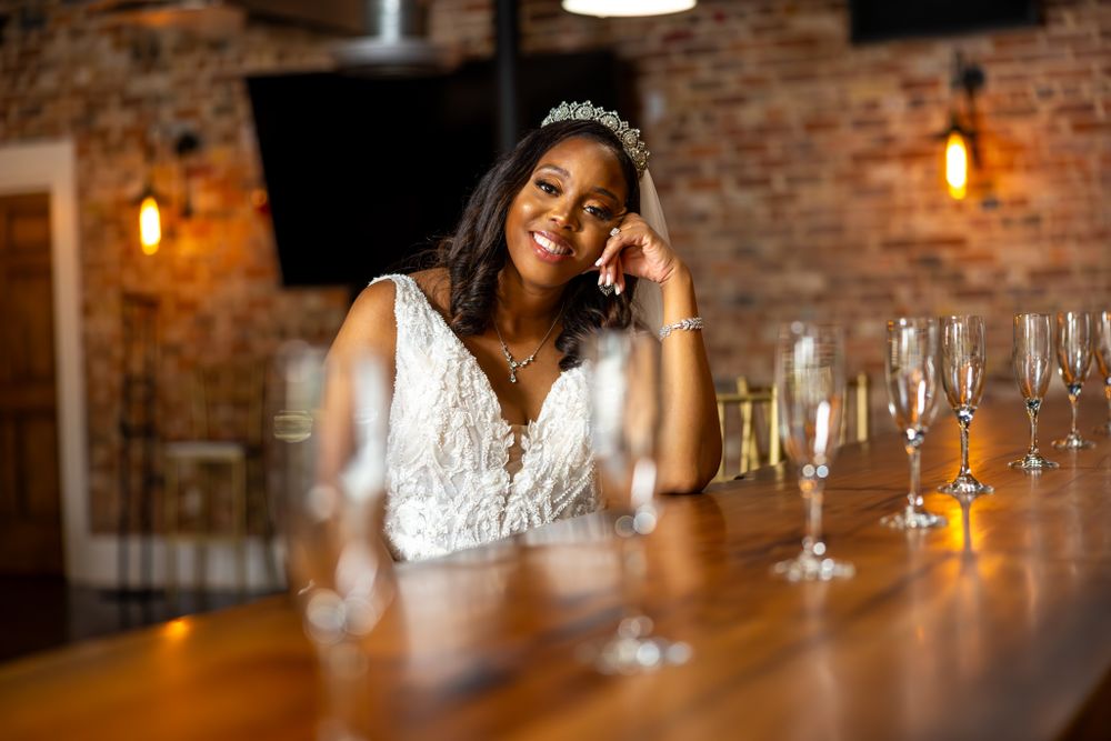 Bride resting at wooden bar with champagne glasses in rustic venue.