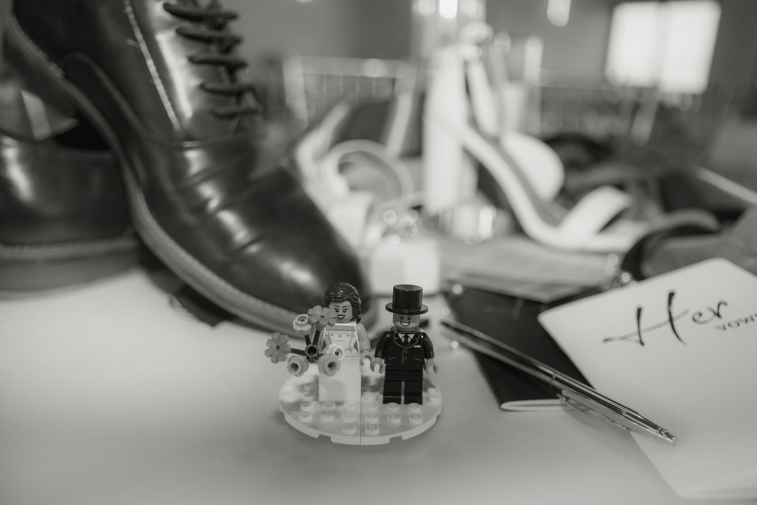 Black and white detail shot of miniature bride and groom figures next to formal wedding shoes.