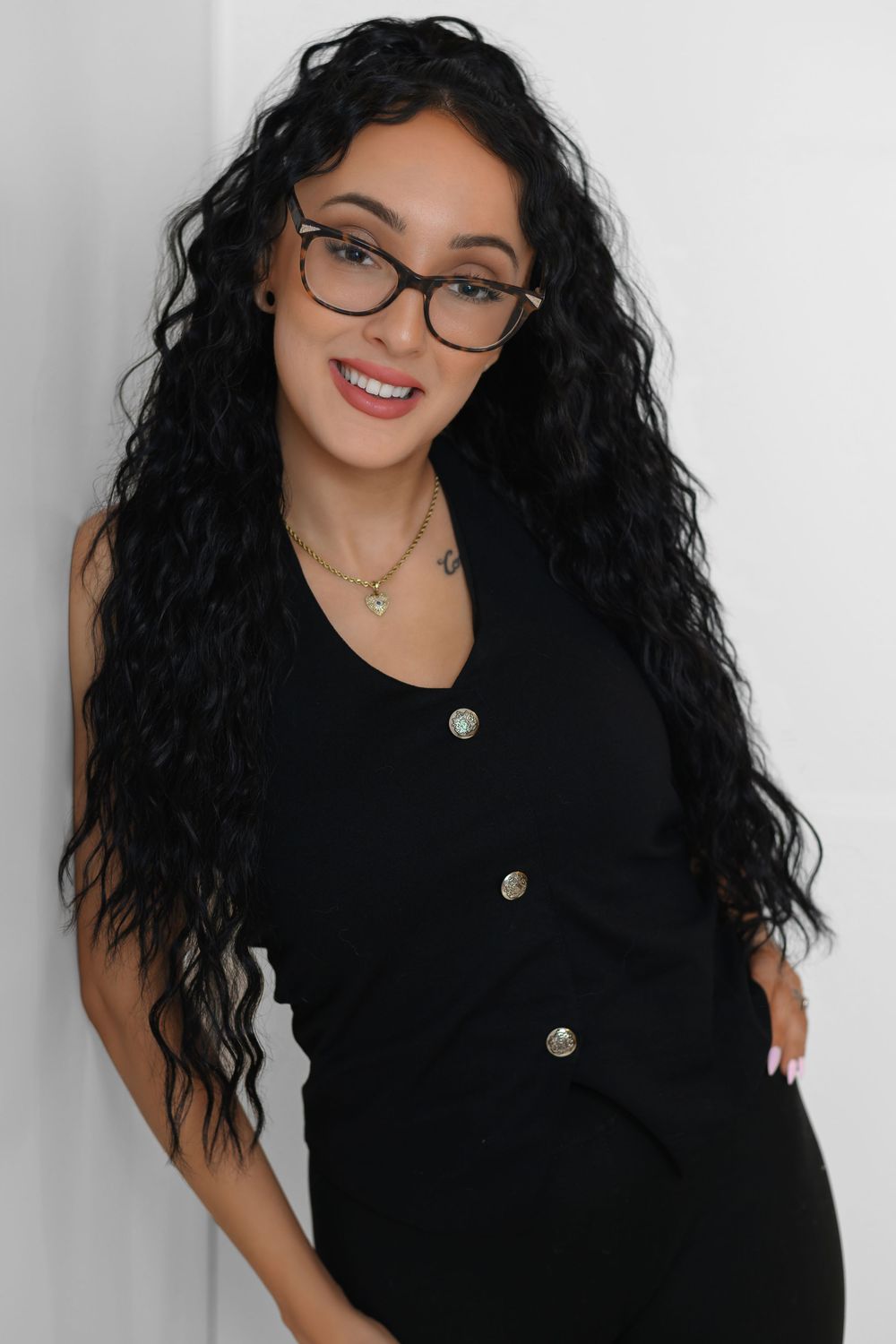 A person with long dark curly hair wearing glasses and a black button-up dress poses against a white wall.