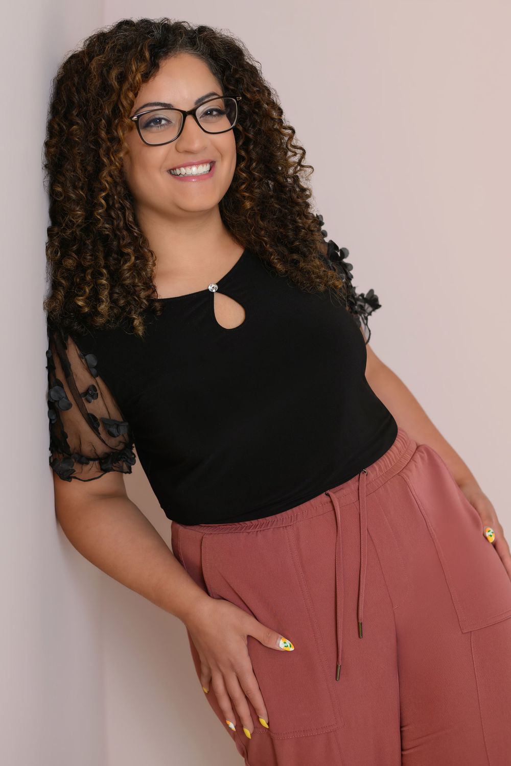 Stylish model in black top and mauve pants poses confidently with curly hair and glasses against light background.