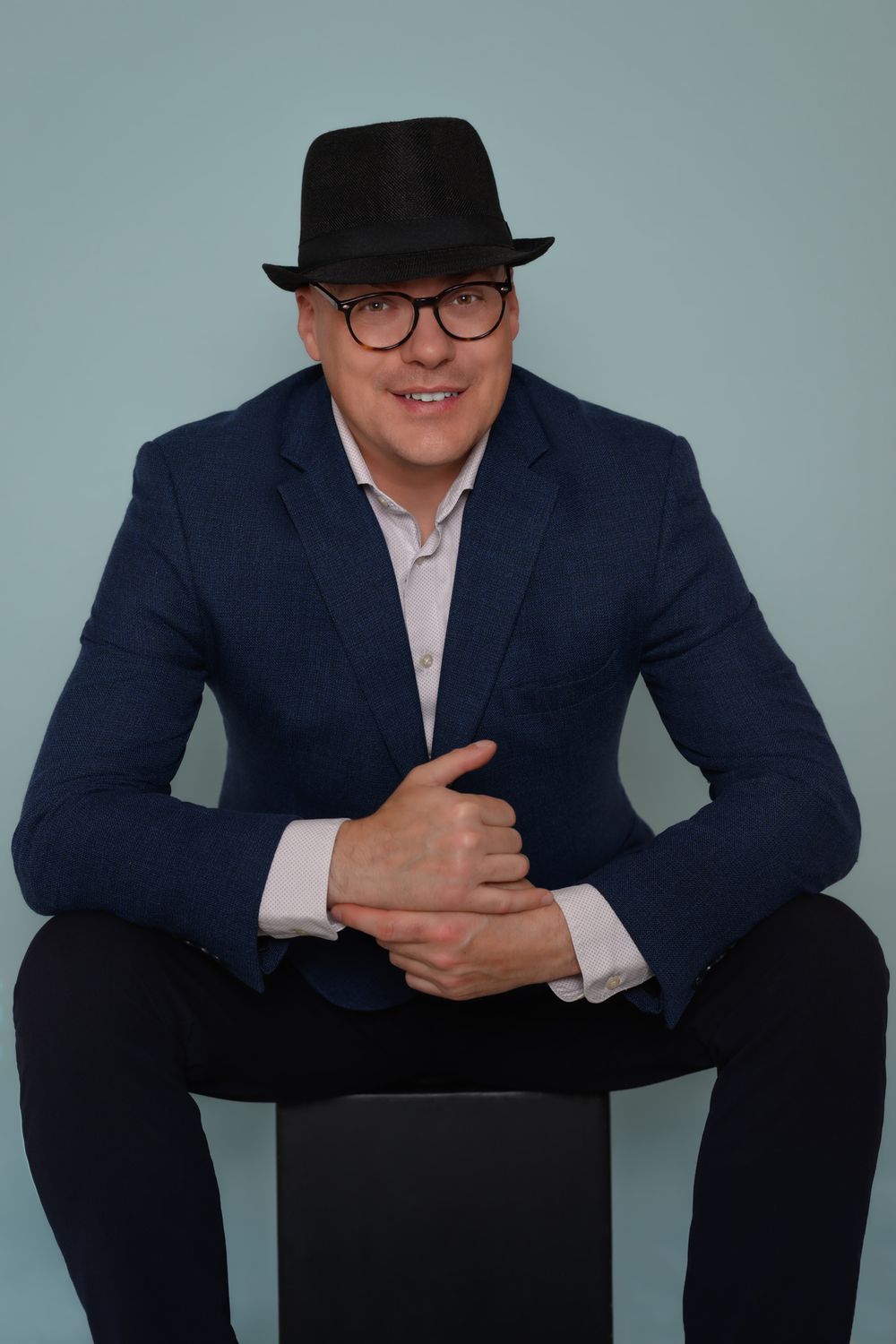Professional wearing navy suit and black fedora sits in various poses against gray studio background.