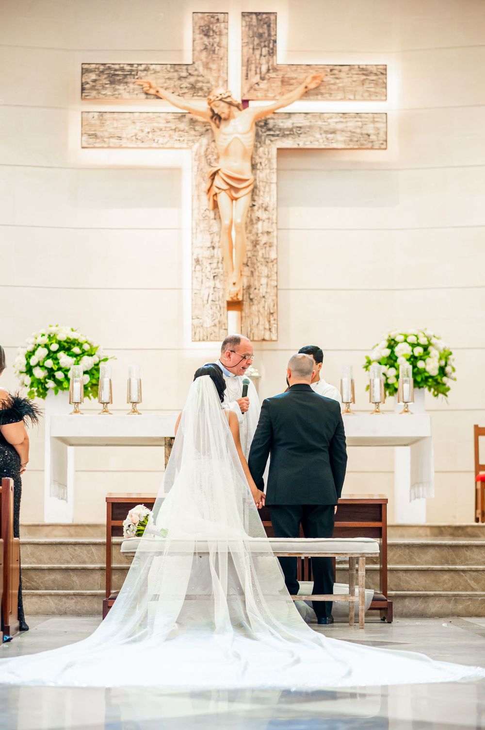 Wedding ceremony at altar beneath large wooden crucifix with floral arrangements.