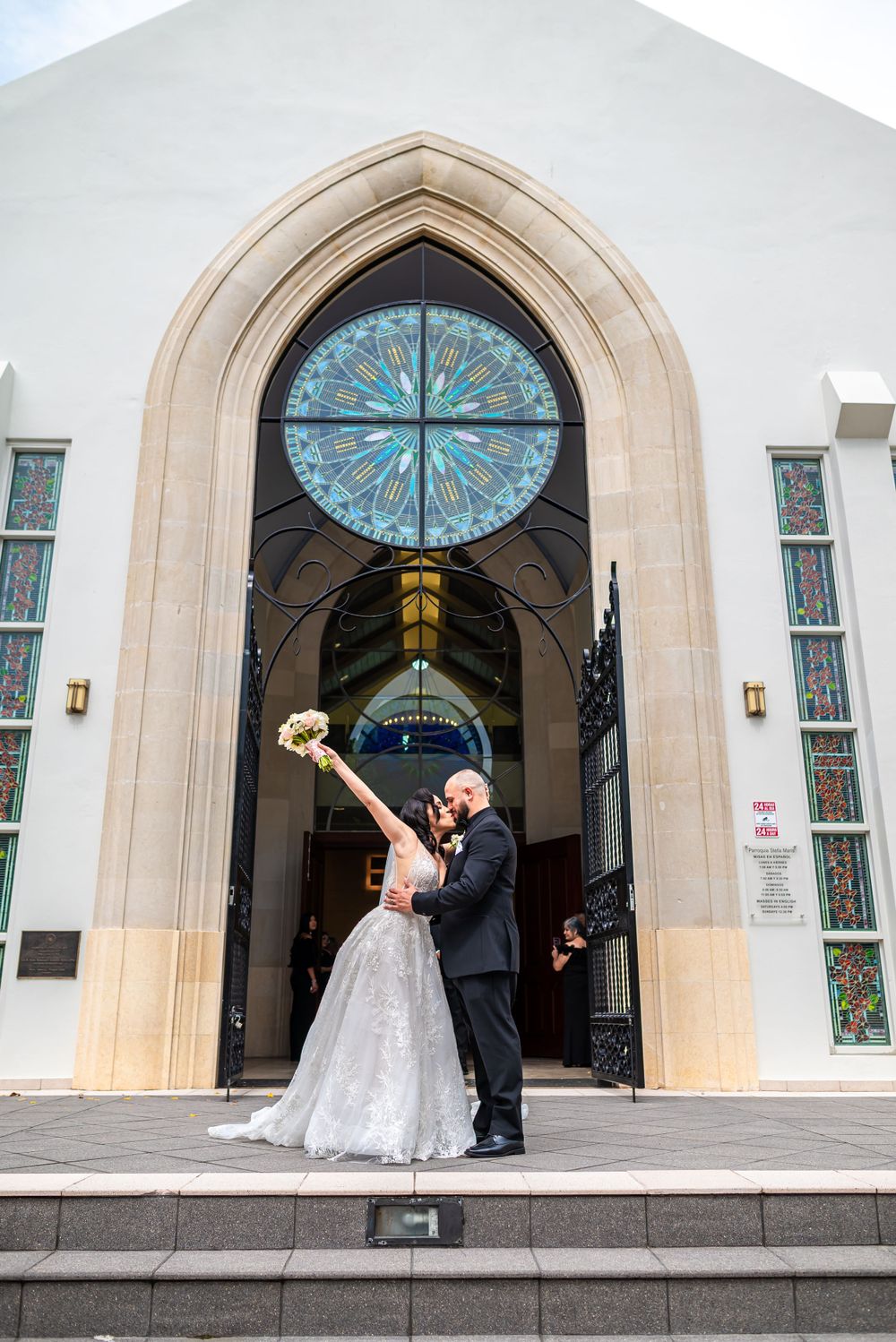 Wedding celebration at church entrance featuring ornate stained glass window and stone archway.