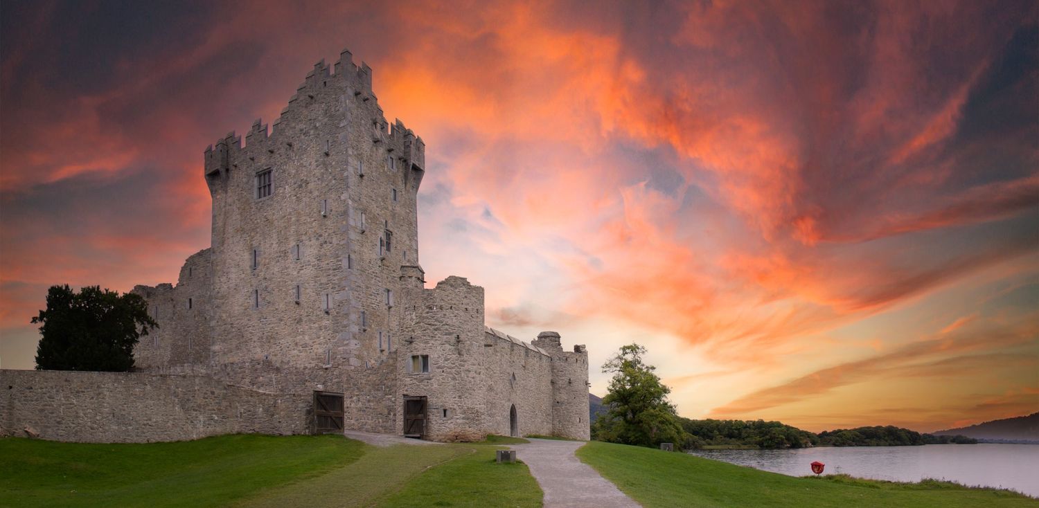Ancient stone castle ruins at dramatic sunset with vibrant red and orange clouds over a calm lake in Ireland.