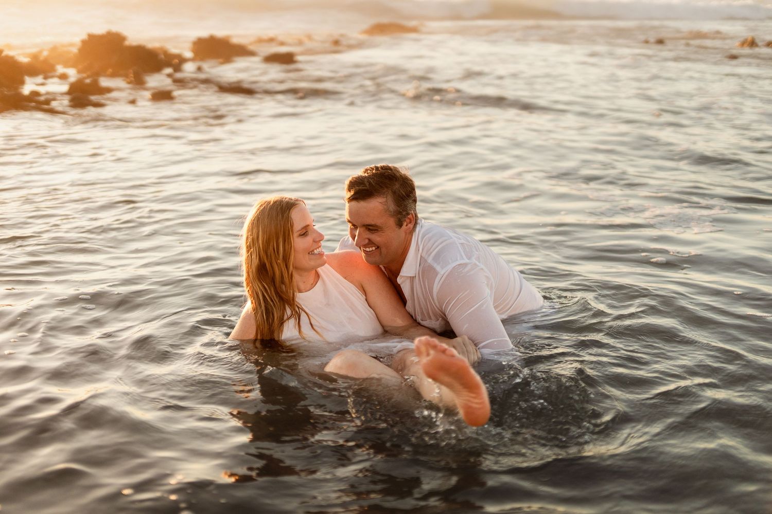 Couple sharing a romantic kiss in ocean waves during a stunning golden sunset at the beach.