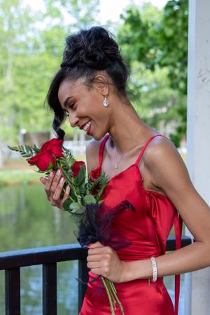 A person in a red dress joyfully smells red roses while standing on a balcony.