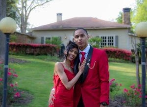 A couple in matching red formal wear pose in a garden with landscaped flowers and lamp posts.