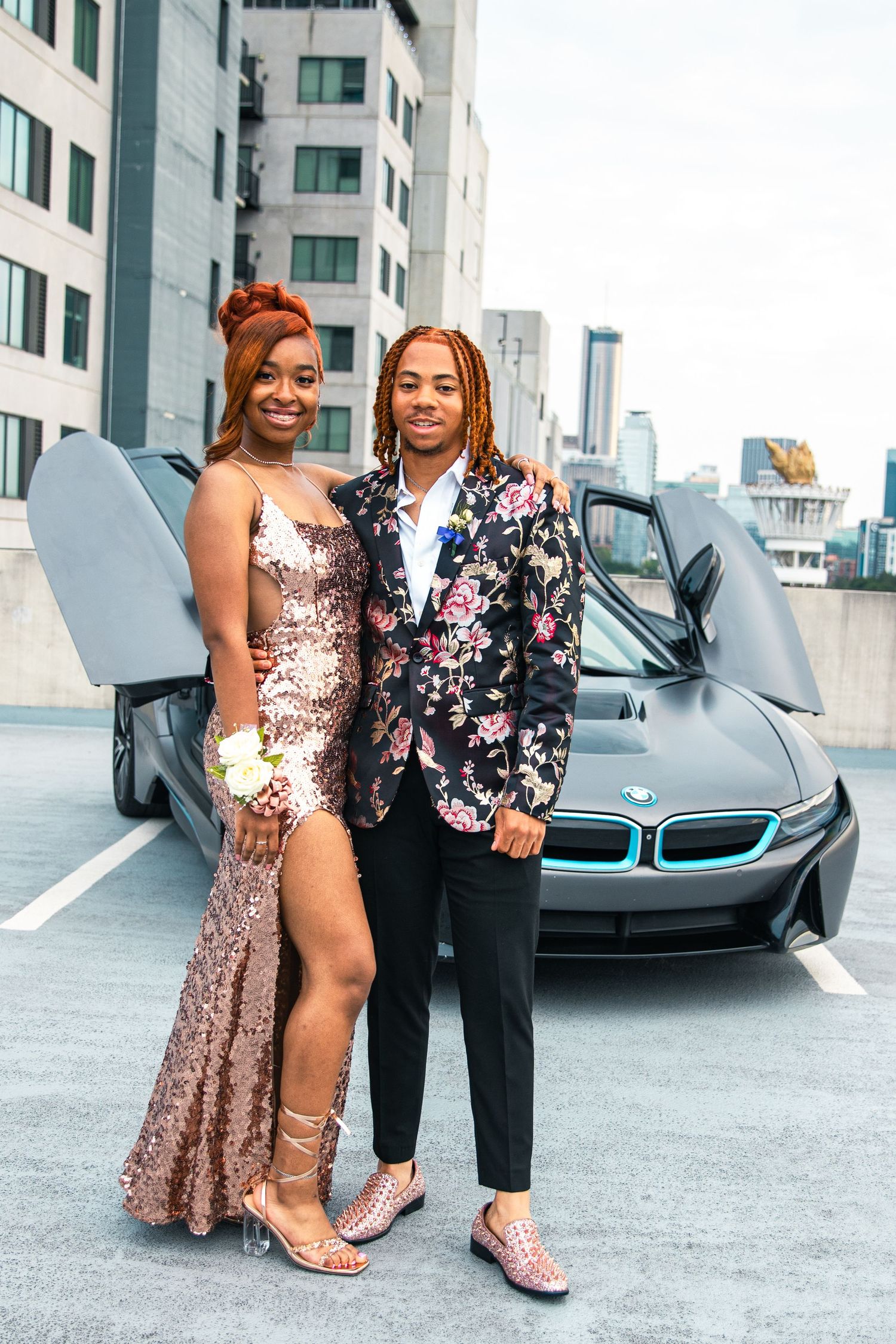 Two people posing in formal attire next to a BMW i8 sports car on a rooftop.