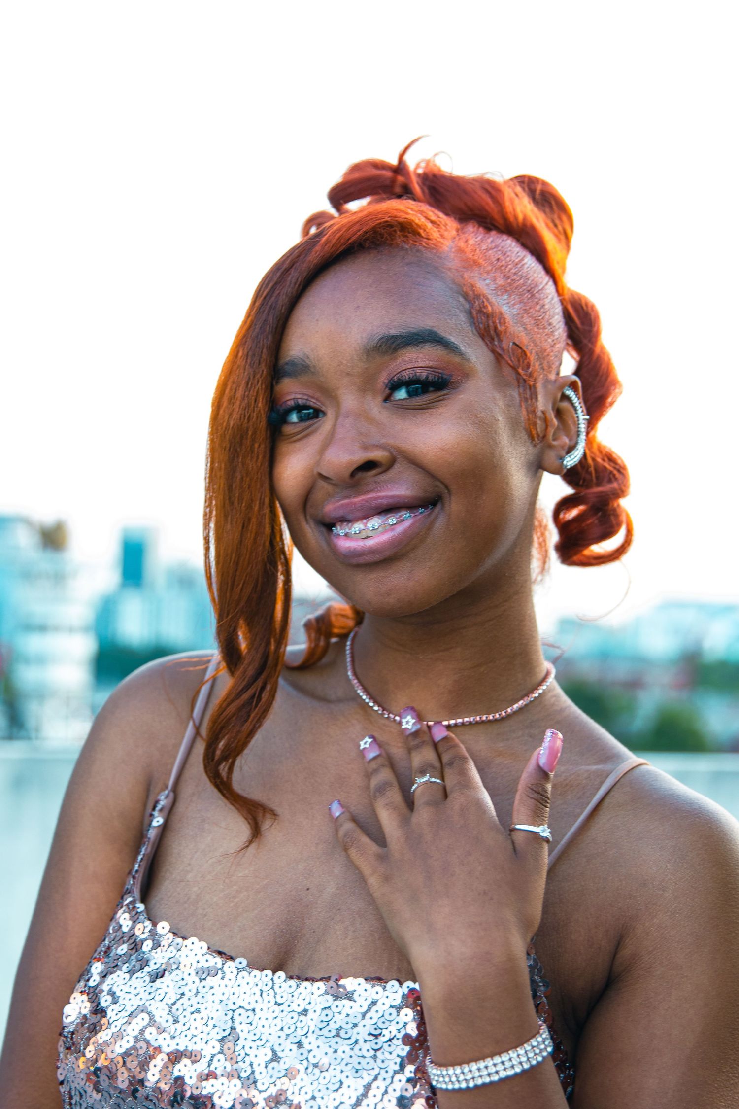 Portrait of person in sequined dress with styled red hair and jewelry against city backdrop.