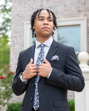 A formal portrait of someone in a black suit with a patterned tie and pocket square against brick building.
