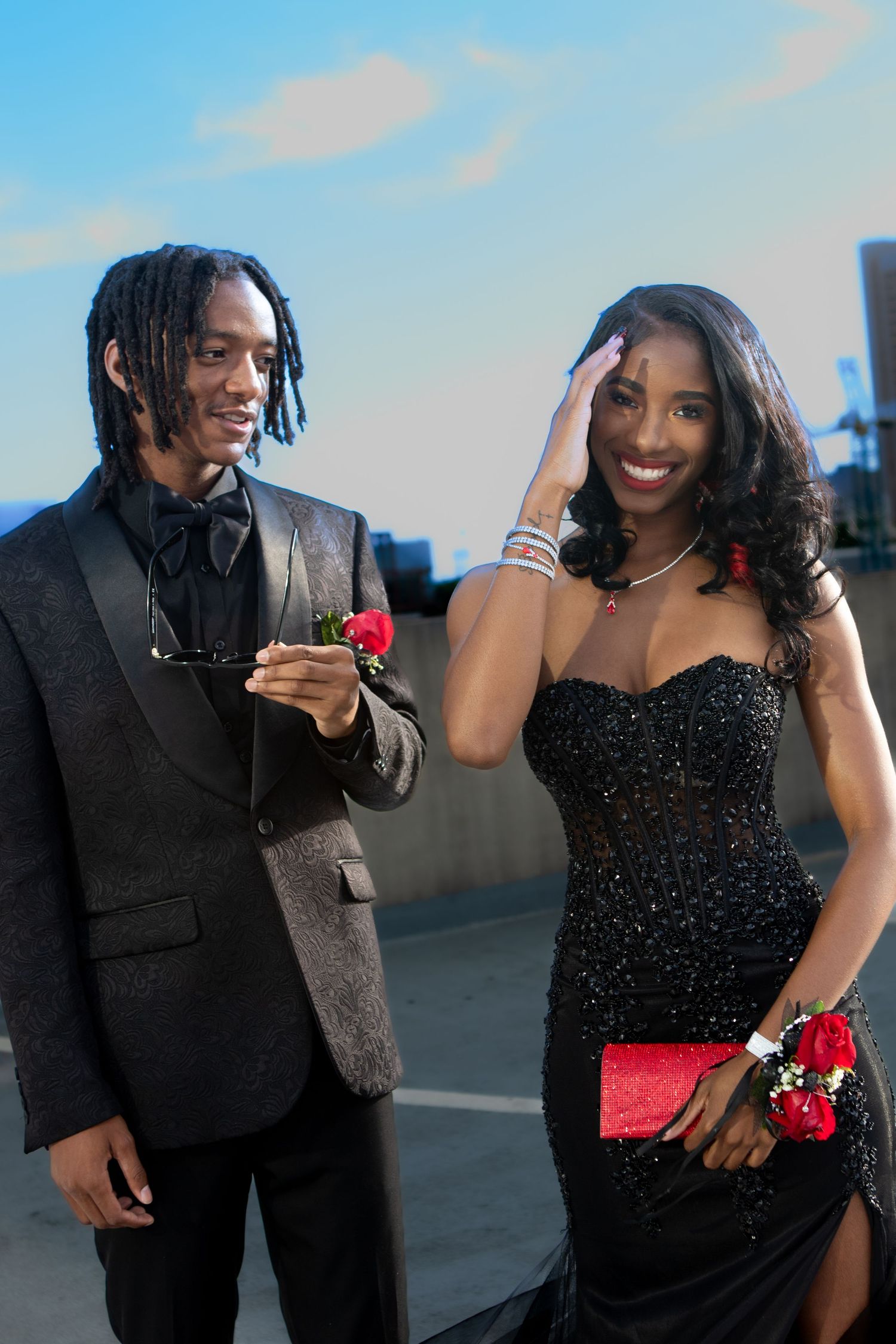 A stylish couple in formal black attire pose outside at prom, with matching red rose corsage and boutonniere.