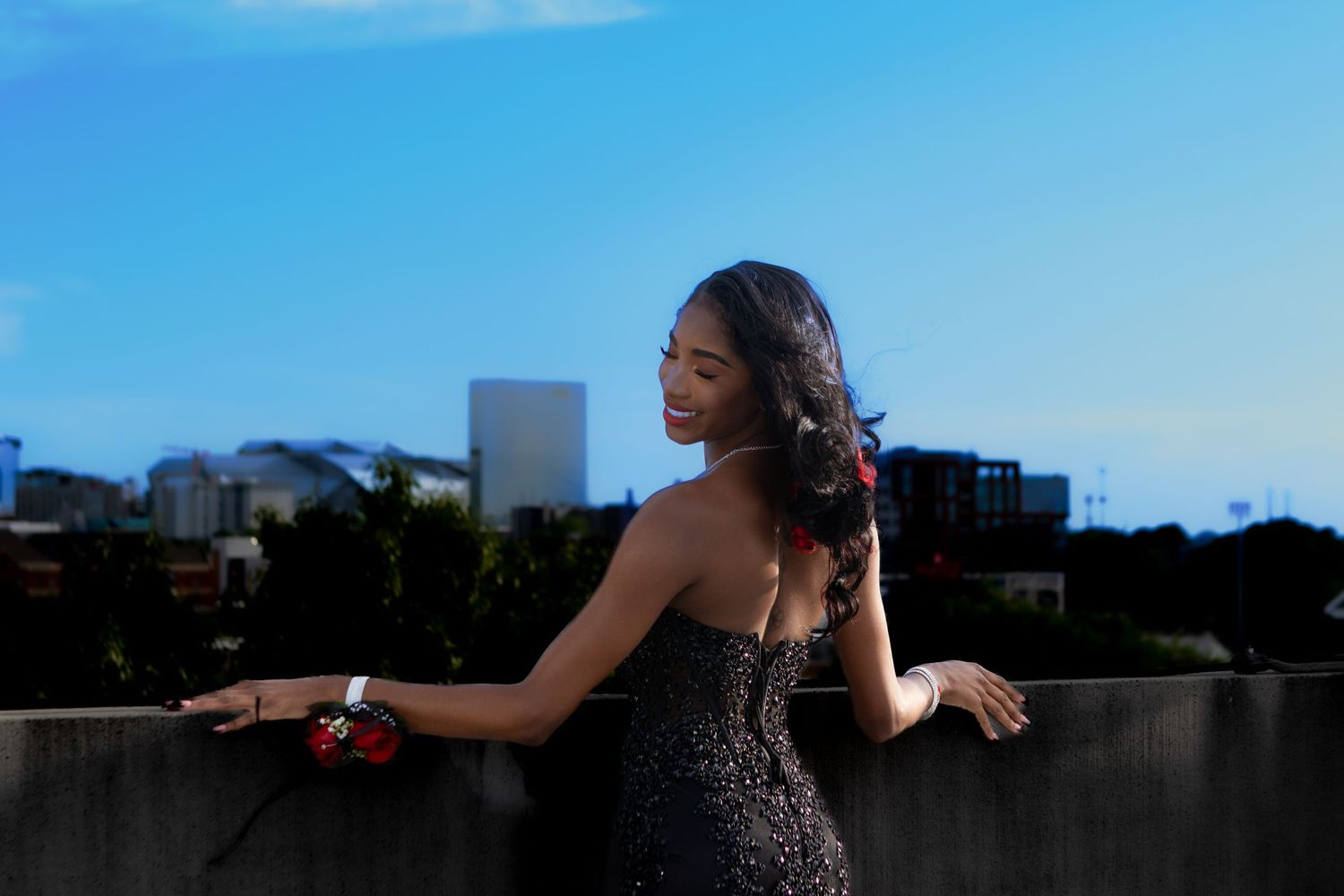 Woman in a sparkly evening dress poses on a rooftop against a city skyline at dusk.