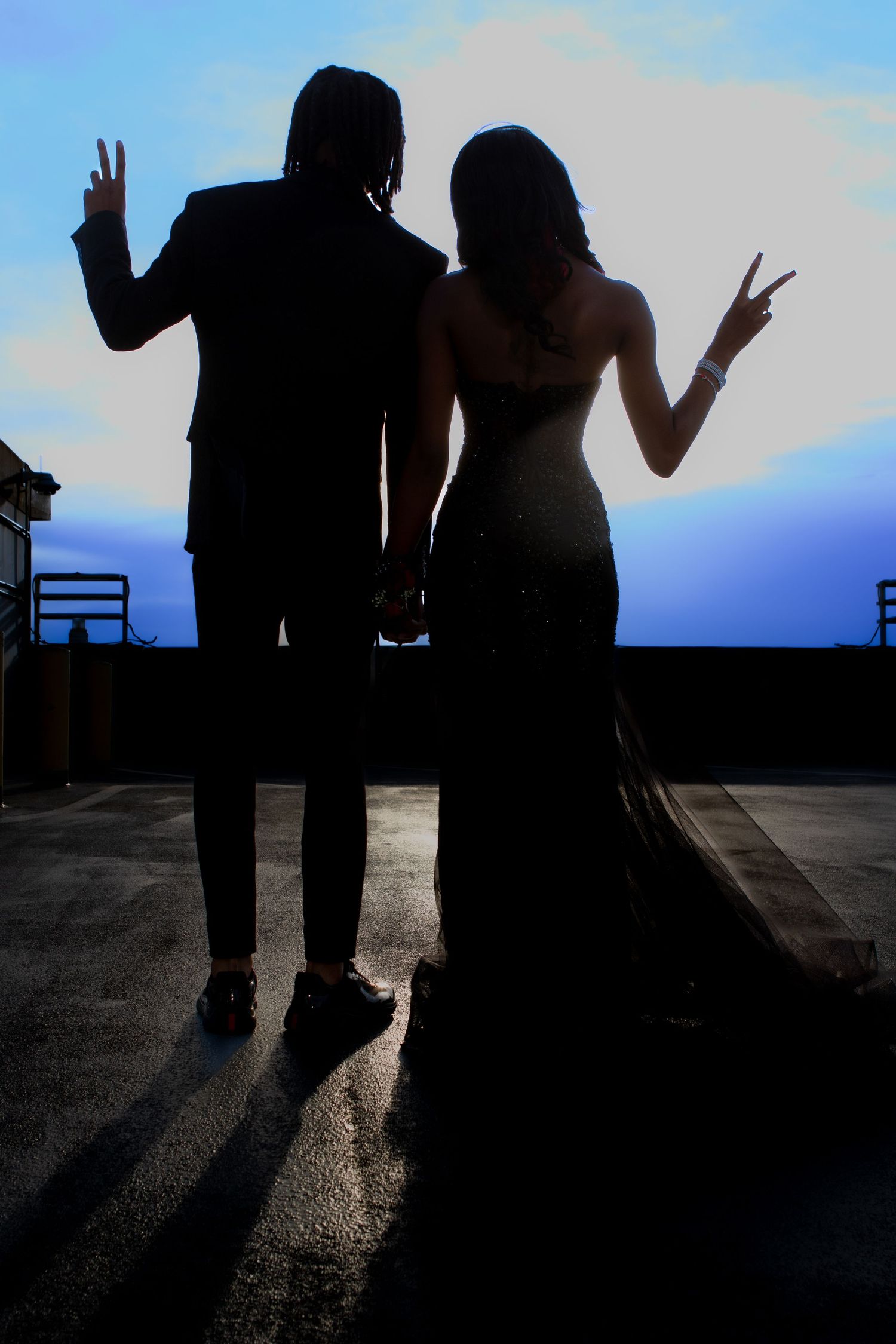 Silhouetted couple making peace signs against a sunset sky on a rooftop with dramatic backlighting.