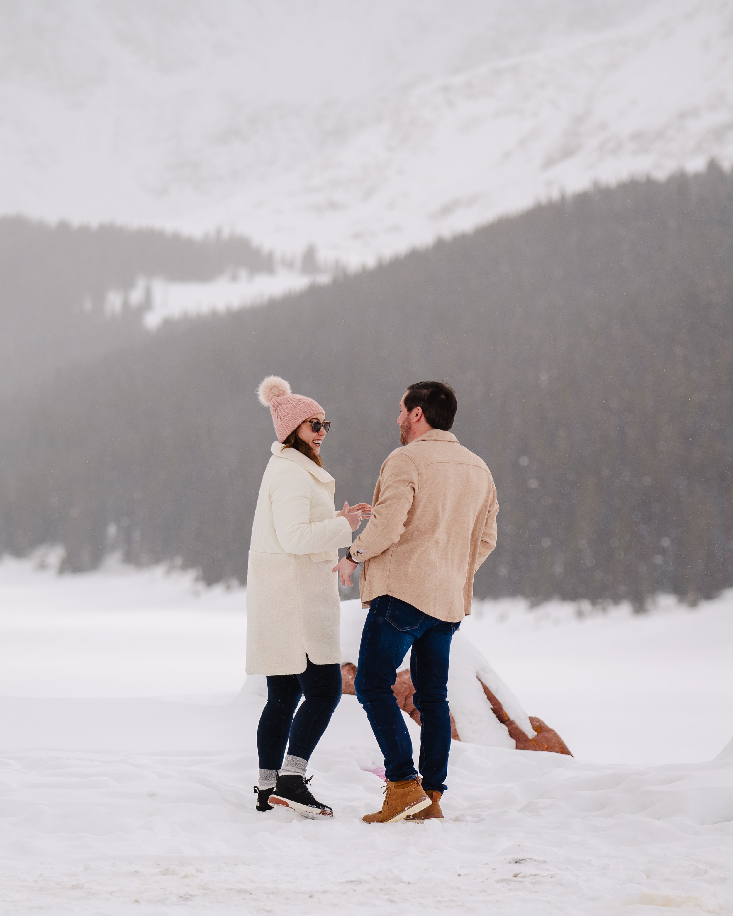 A couple stands together in snowy winter landscape with mountain backdrop wearing warm coats.