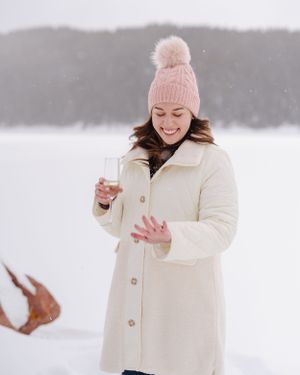 Person in cream coat and pink beanie looking down at ring in snowy setting.