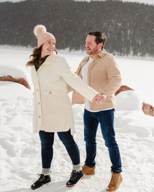 A playful winter scene with a couple dancing together in the snow wearing coordinating coats.