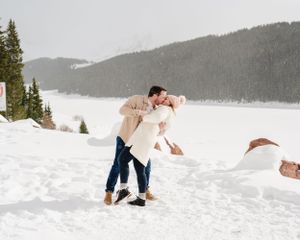 A romantic winter scene showing a couple dancing in the snow near a mountainside.