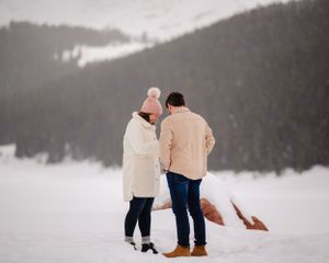 Two people in coordinating winter outfits stand together in a snowy mountain setting.