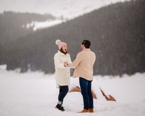 Two people share a tender moment while kneeling in fresh snow with evergreen mountains behind them.