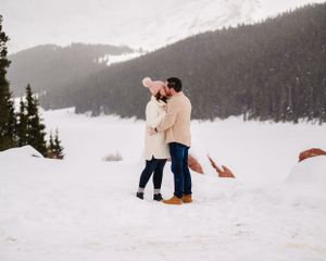 Two people share an emotional moment in deep snow with evergreen forests and mountains in view.