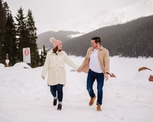 Two people embrace joyfully in deep snow with evergreen trees and mountains behind them.