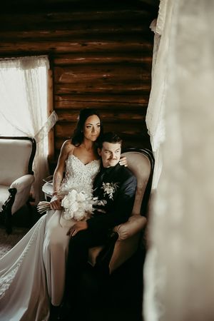A couple in wedding attire sits together in an armchair inside a rustic log cabin with dramatic moody lighting.