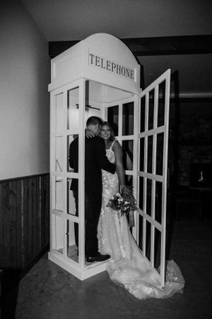 Black and white image of a couple posing in a white decorative telephone booth, with a flowing wedding dress train.