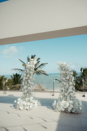 Two white floral arch columns frame a stunning beachfront wedding ceremony venue with palm trees and turquoise ocean views.