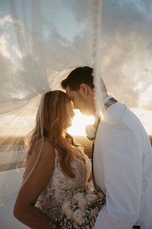 Bride and groom share romantic moment under flowing veil at sunset, bride holding white bouquet in lace wedding dress.