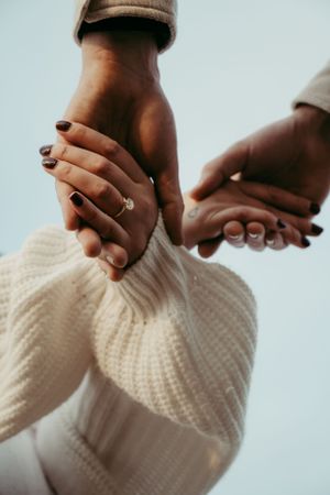 Two hands clasped together showing an engagement ring, with one person wearing a cozy white knit sweater.