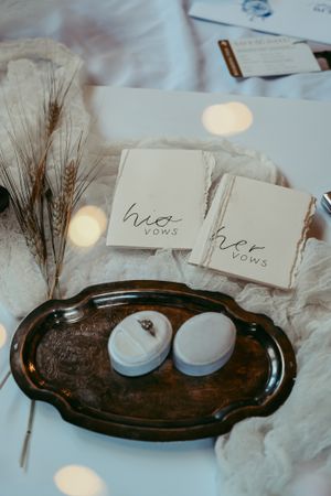 Wedding vow books and ring boxes displayed on vintage silver tray with dried wheat stalks and soft bokeh lighting.