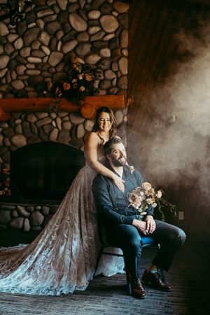 Couple in romantic indoor setting with stone fireplace, displaying elegant dress and bouquet in moody atmosphere.