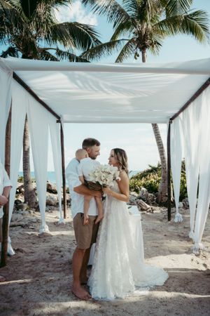 A couple shares a romantic moment under a white canopy on a tropical beach with palm trees in the background.