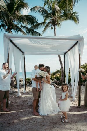 A desintation beach wedding ceremony under a white canopy draped with fabric alongside palm trees on a tropical shoreline.