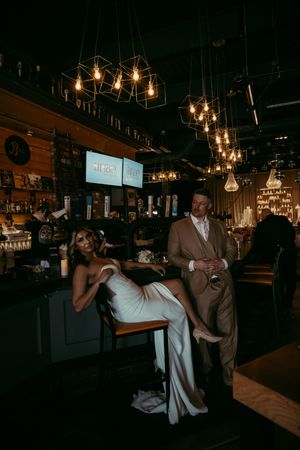 Bride in white gown sits at bar while groom in tan suit stands beside her, surrounded by warm Edison bulb lighting.