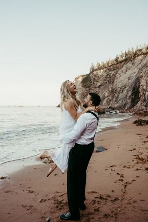 A couple embraces on a scenic beach with rocky cliffs and ocean waves at sunset.