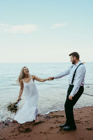 A couple shares a romantic moment on the beach, holding hands while dressed in wedding attire against the ocean backdrop.