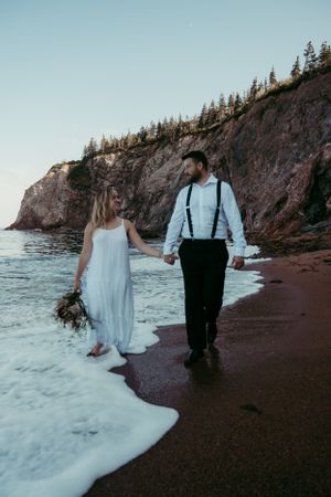 A couple walks along a dramatic rocky beach at sunset with towering cliffs and crashing waves in Nova Scotia.