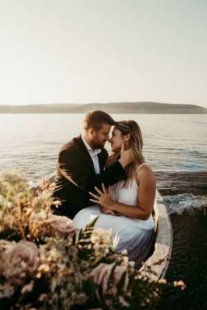 A romantic couple embraces at sunset by the ocean with pink flowers in the foreground.