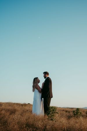 A couple in wedding attire stands silhouetted against a blue sky on a hilltop covered in golden grass at sunset.