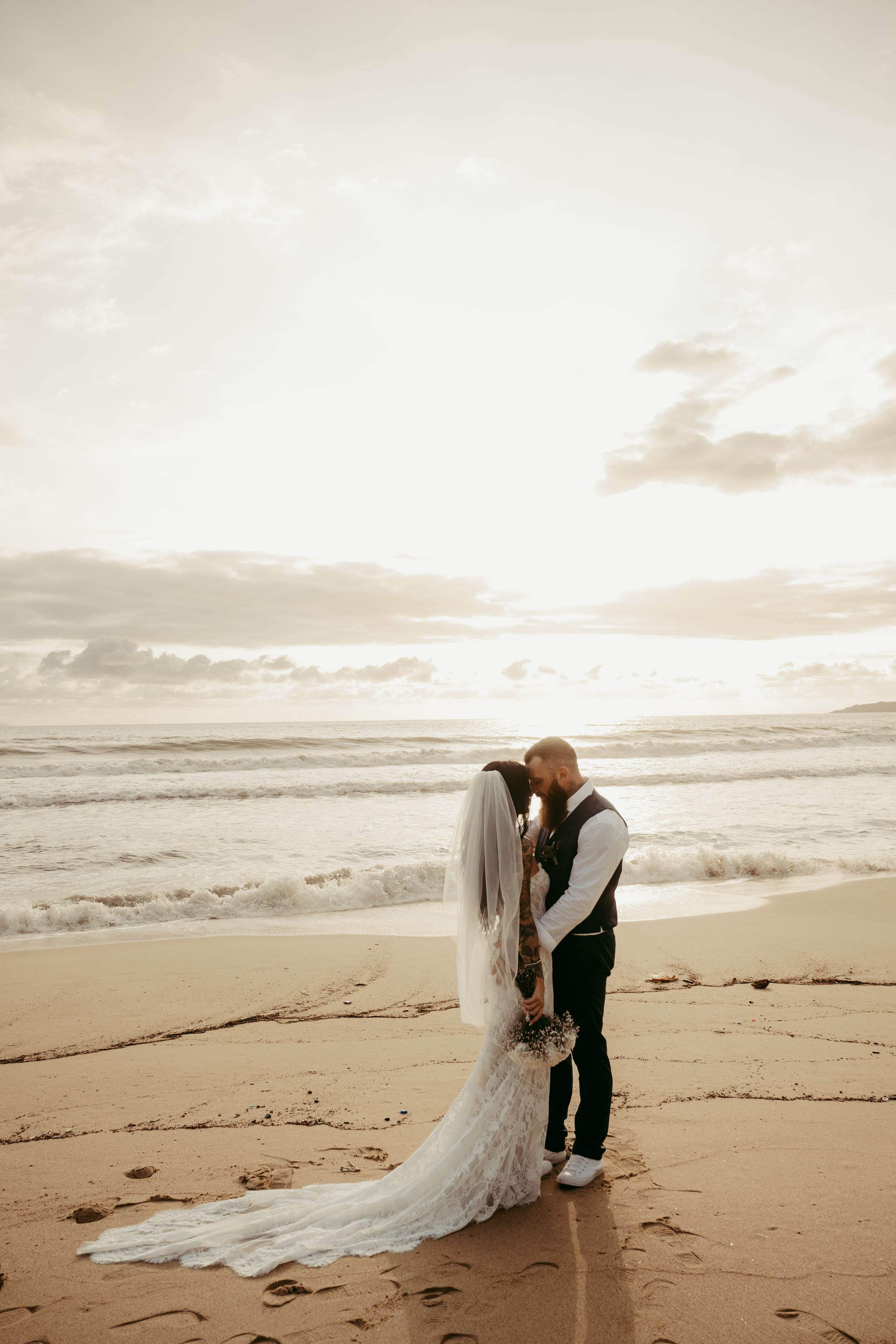 Bride and groom share romantic beach moment at sunset, waves crashing behind them, wedding dress trailing on sand.