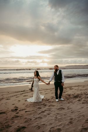 Bride and groom walk hand-in-hand on a sandy beach at sunset, waves crashing behind them on their wedding day.
