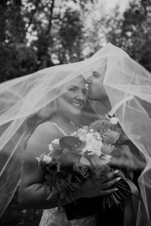 A romantic black and white wedding photo showing a couple embracing under a flowing bridal veil with a floral bouquet.