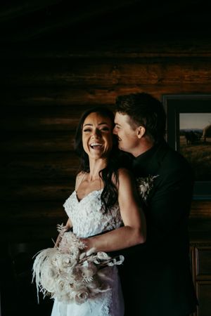 A couple in wedding attire share a candid moment in front of a rustic wooden wall.