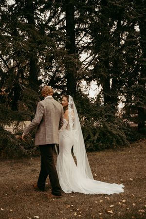 A bride in a white lace gown and veil walks with a groom in a tan suit among tall evergreen trees.