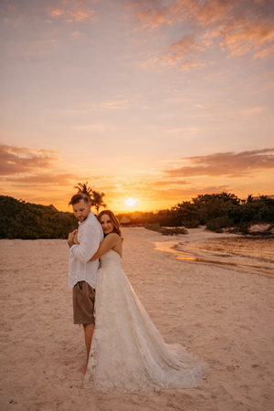 Couple embracing on sandy beach at golden sunset, bride in lace gown, groom in white shirt, tropical trees backdrop.
